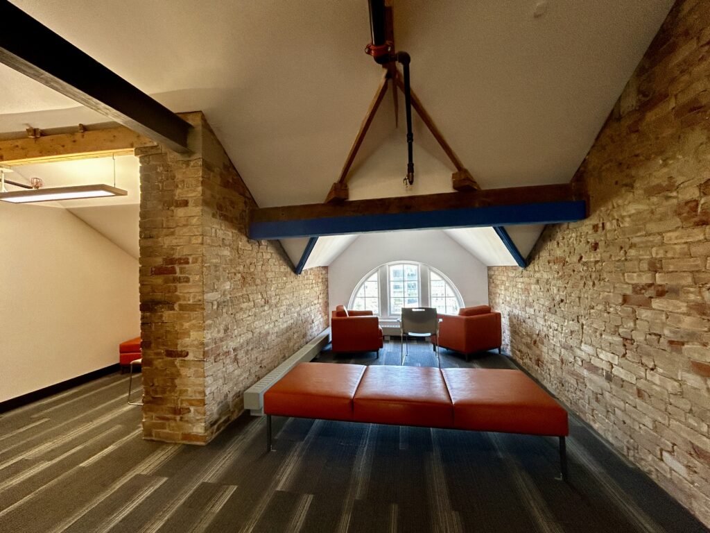 A wide shot of the attic at G building which features a couple of orange couches. The are has low white ceiling, brick walls, and carpeted floors.