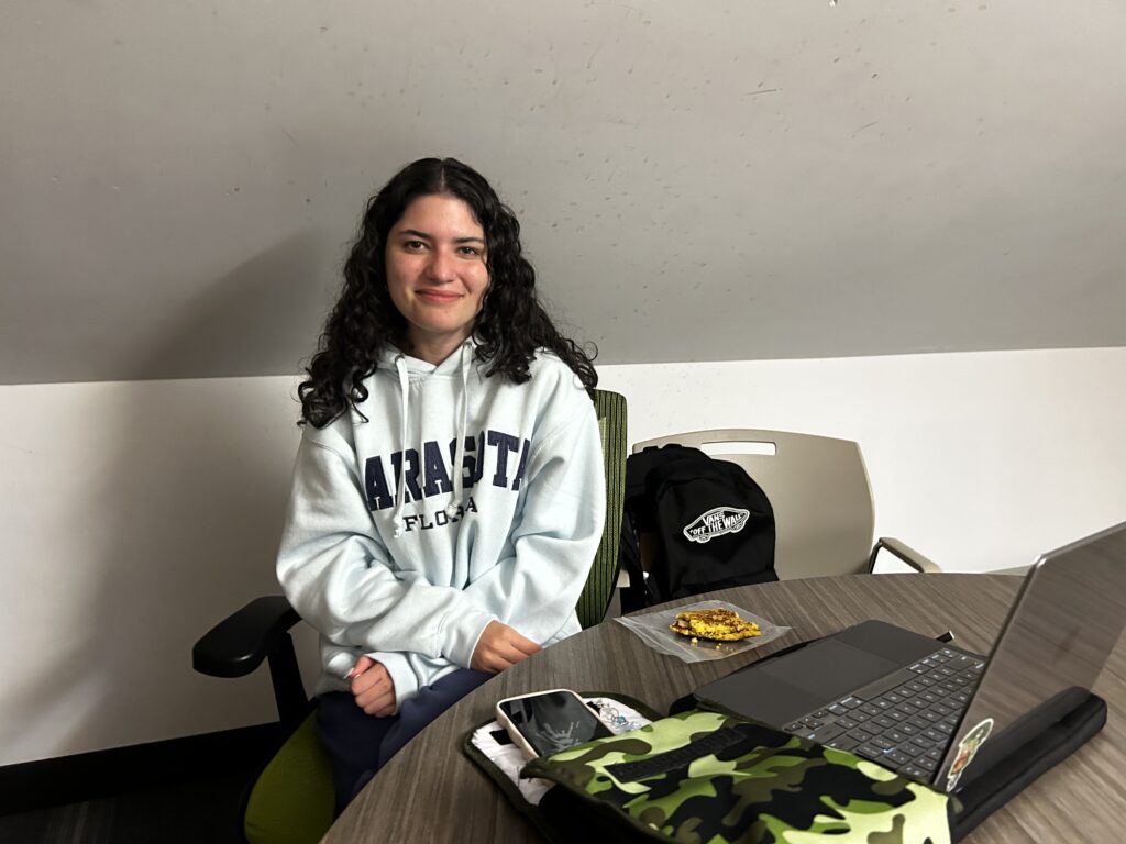 A girl in a white hoodie sits behind a table containing her laptop, phone, bag, and a cookie. 