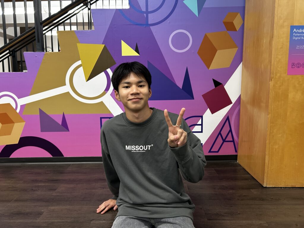 A boy smiles as he raises his left hand in a peace sign. His background is a mural found in the basement area of the F building.