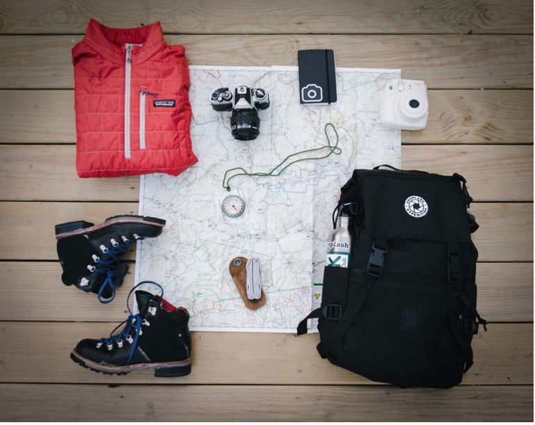 Black hiking backpack near white Fujifilm instax mini camera near black leather boots, red half-zip jacket, gray pocket watch on white map.