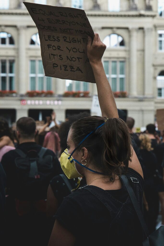 A girlr wearing a yellow face mask holds up a cardboard sign that reads, "Equal rights 4 everyone does NOT mean less rights for you, it's not a pizza".