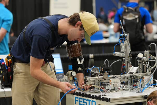 Cole Hunter with a brown bracer and tool belt, bending over to work on wiring of a complex system.