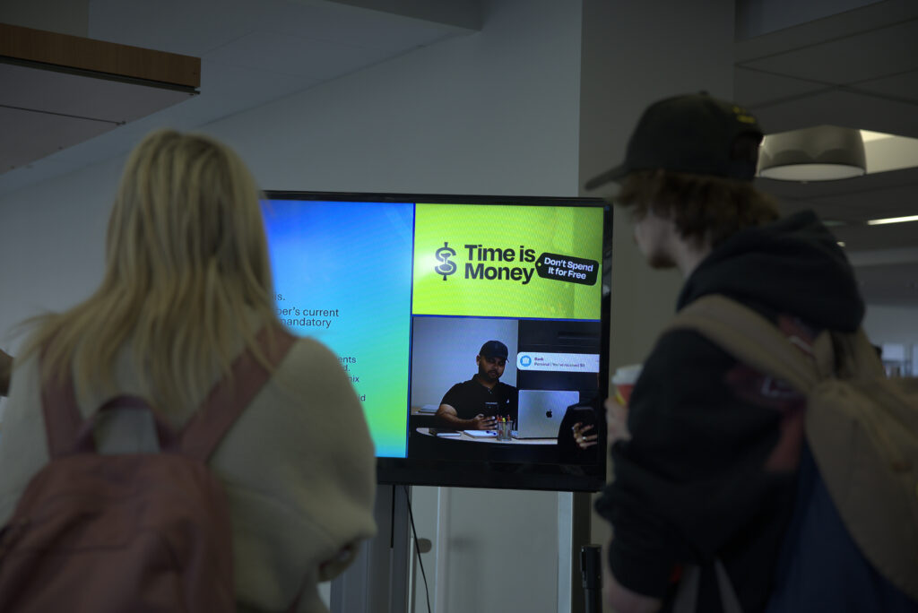 Two students are staring at a TV with a video of IGNITE's Time is Money campaign.