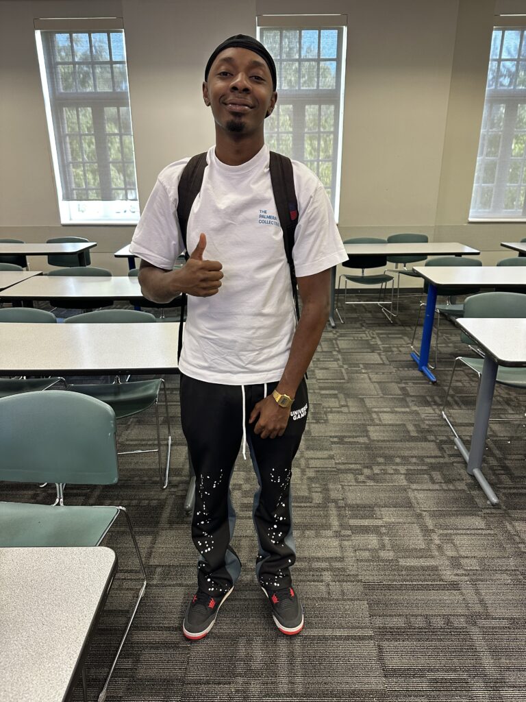 Student in a white t-shirt, and skinny jeans, in a classroom, giving a thumbs up sign