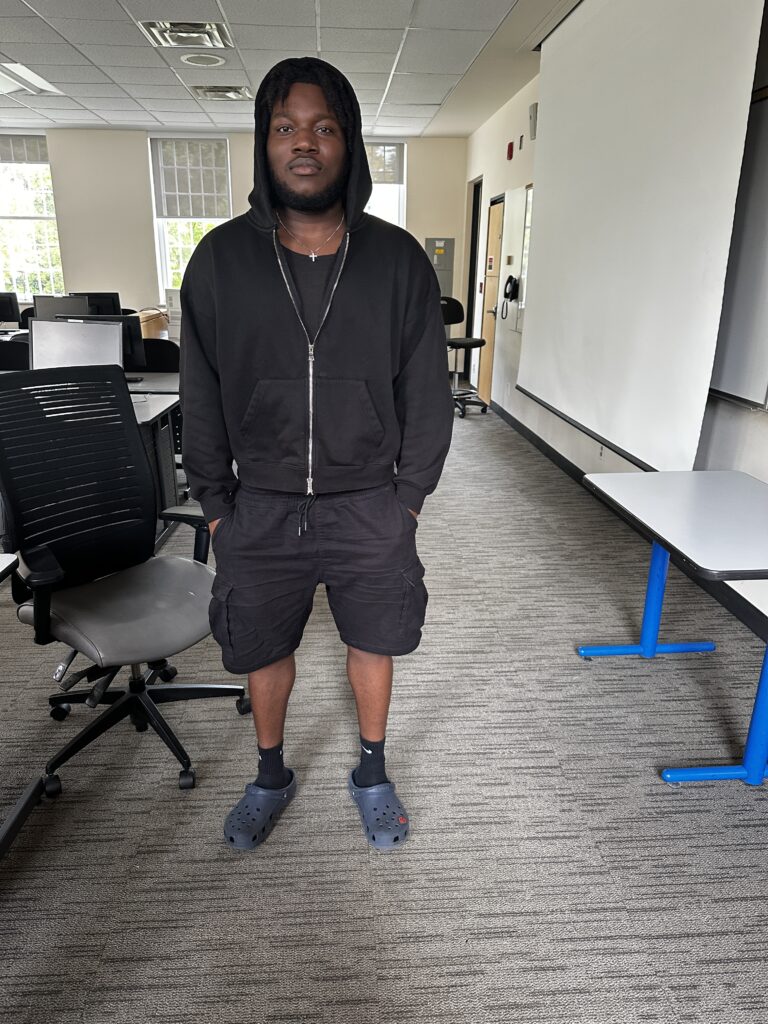 Student in a black hoodie, and black shorts, in a classroom, with his hands in his pockets facing forward.
