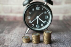 An black analong clock behind three stacks of bronze coins.