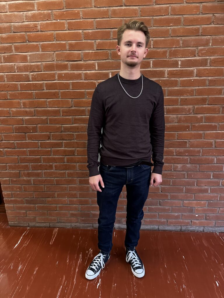 Image of a student looking into the camera for a photo, with a silver chain on and the background of red and brownish bricks.