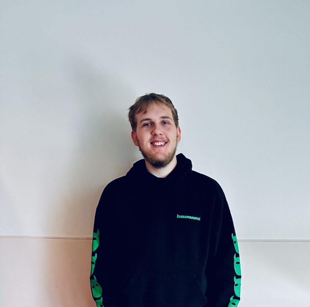 Image of a student looking into the camera for a photo, he is smiling and has a black and green hoodie on.