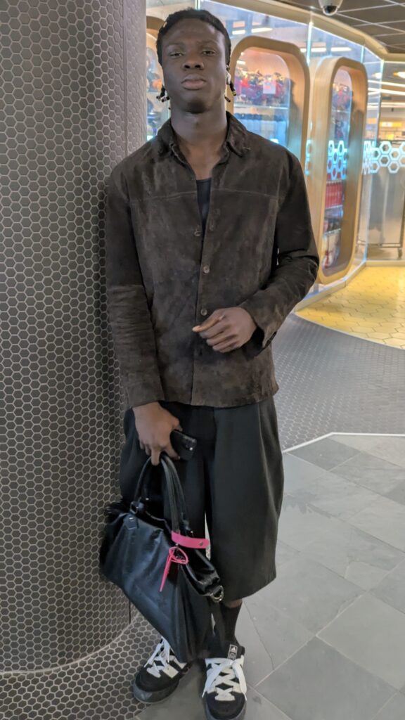 Amani Akinwami wearing a brown jacket, capri pants and holding a handbag looking sternly at the camera.