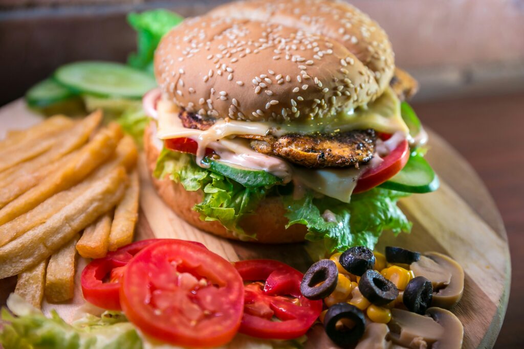 A meal consisting of a burger with a slew of topics and sauce oozing out the side. French fries, tomatoes, olives, mushrooms and corn on a wooden circular plate.