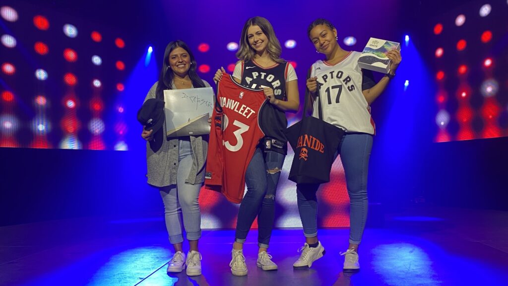 Three women wearing Toronto Raptors gear and posing for a picture on a stage.