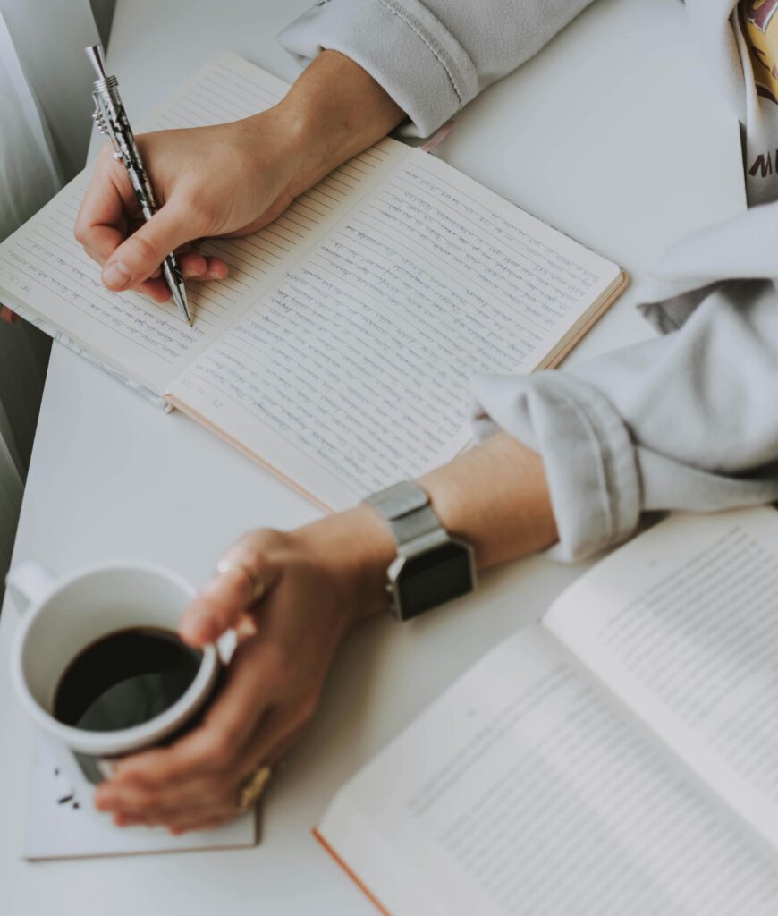 A person's hand writing in a notebook while the other hand holds a cup of coffee. 
