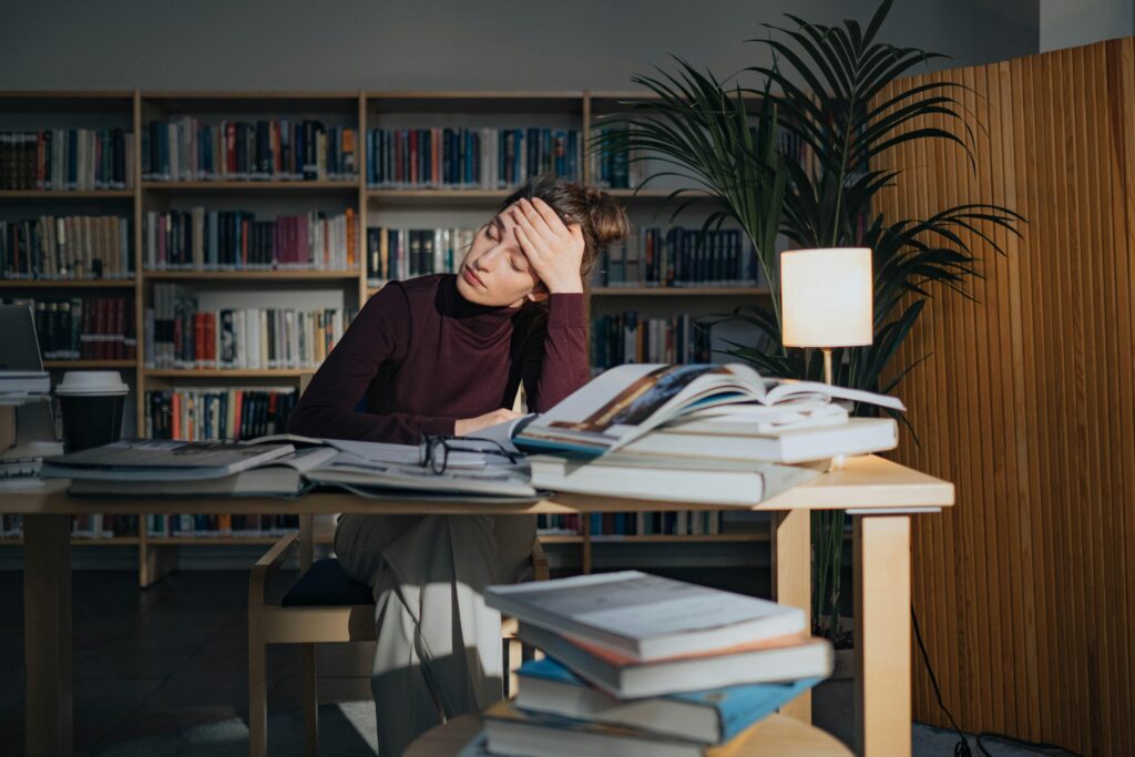 A person holding their hand to their forehead while sitting at a table full off half open textbooks.