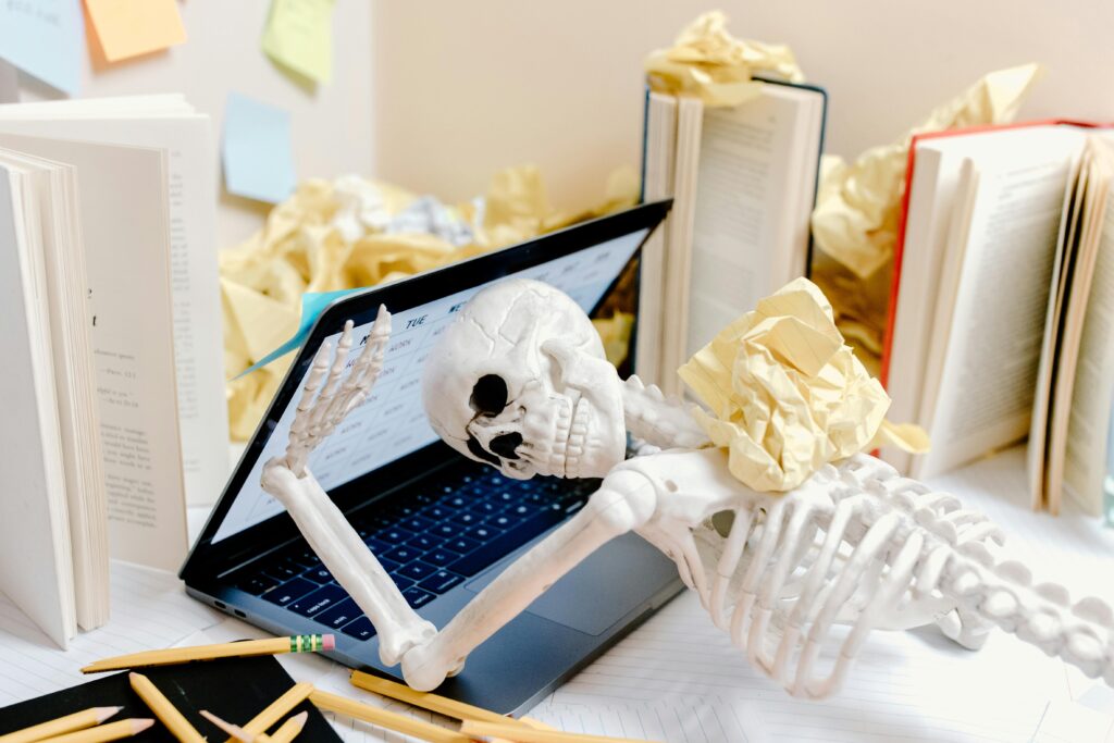 a skeleton leaning over an open laptop holding the laptop open surrounded by books. 