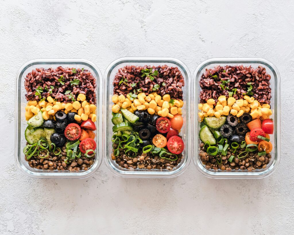 Three glass containers filled with layered sliced cucumbers, cherry tomatoes, corn, olives and red rice, arranged neatly on a light surface.
