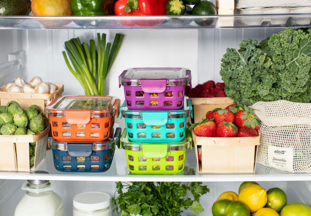 Organized refrigerator shelf with assorted vegetables, fruits, and stackable multicoloured food storage containers.