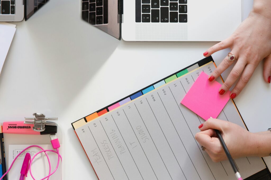 A person writes on a pink sticky note beside an open planner with labelled tabs and two laptop keyboards visible on a white desk.
