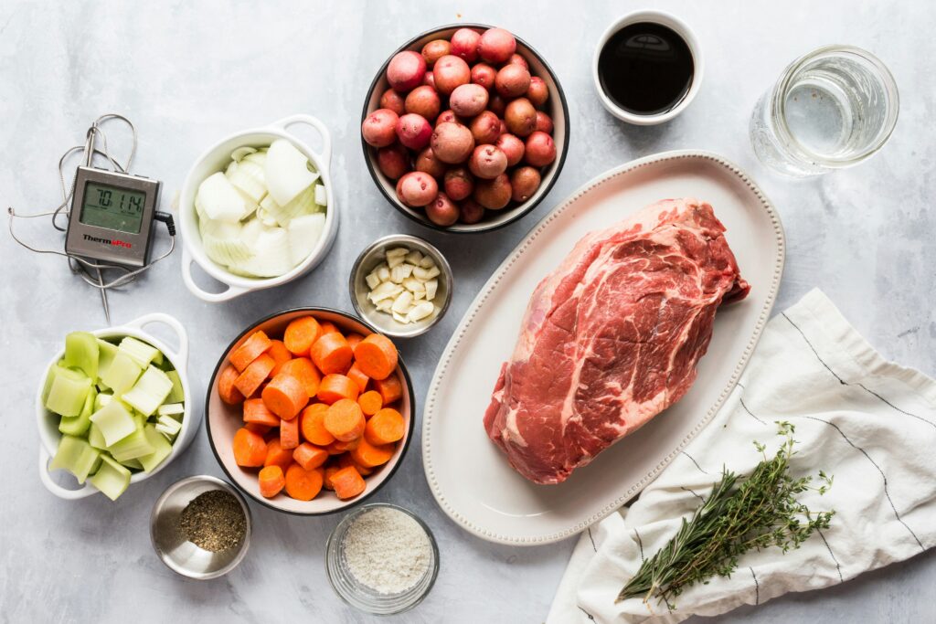 Raw ingredients for a pot roast are arranged on a table, including a beef roast, potatoes, carrots, onions, garlic, spices, broth, water and a digital thermometer.