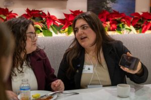 Two women seated at a table talk as one shows something on her phone.