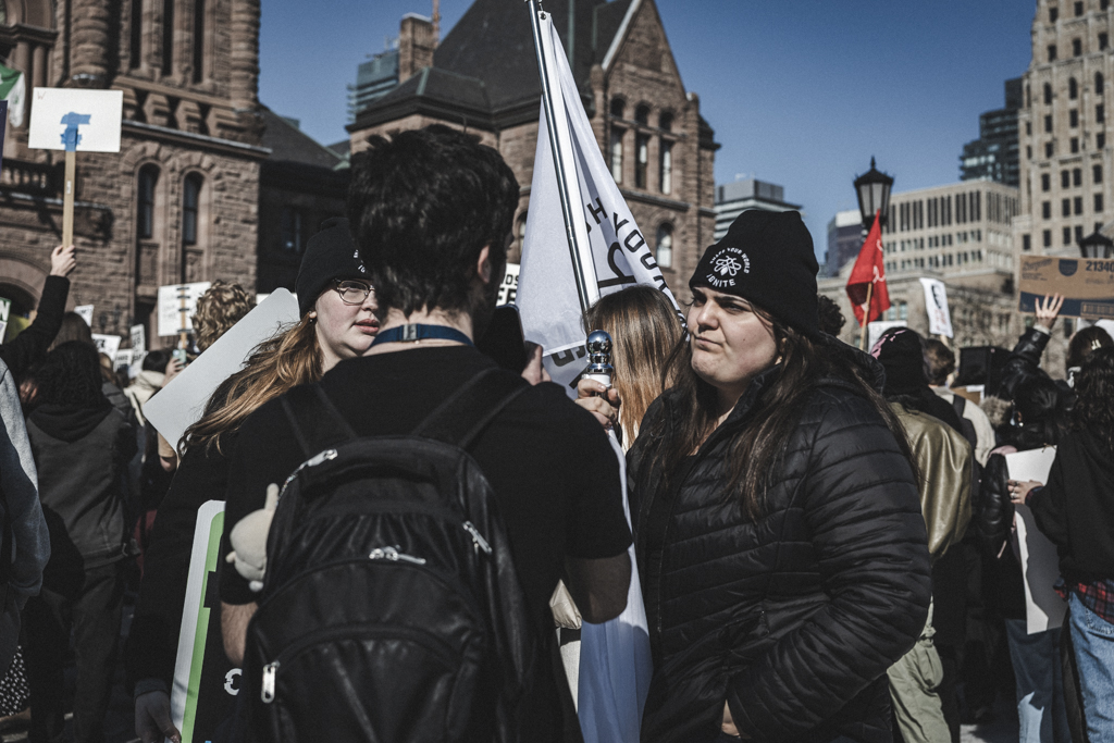 Two students are being interviewed during a rally, with people in the background holding banners, signs and flags.