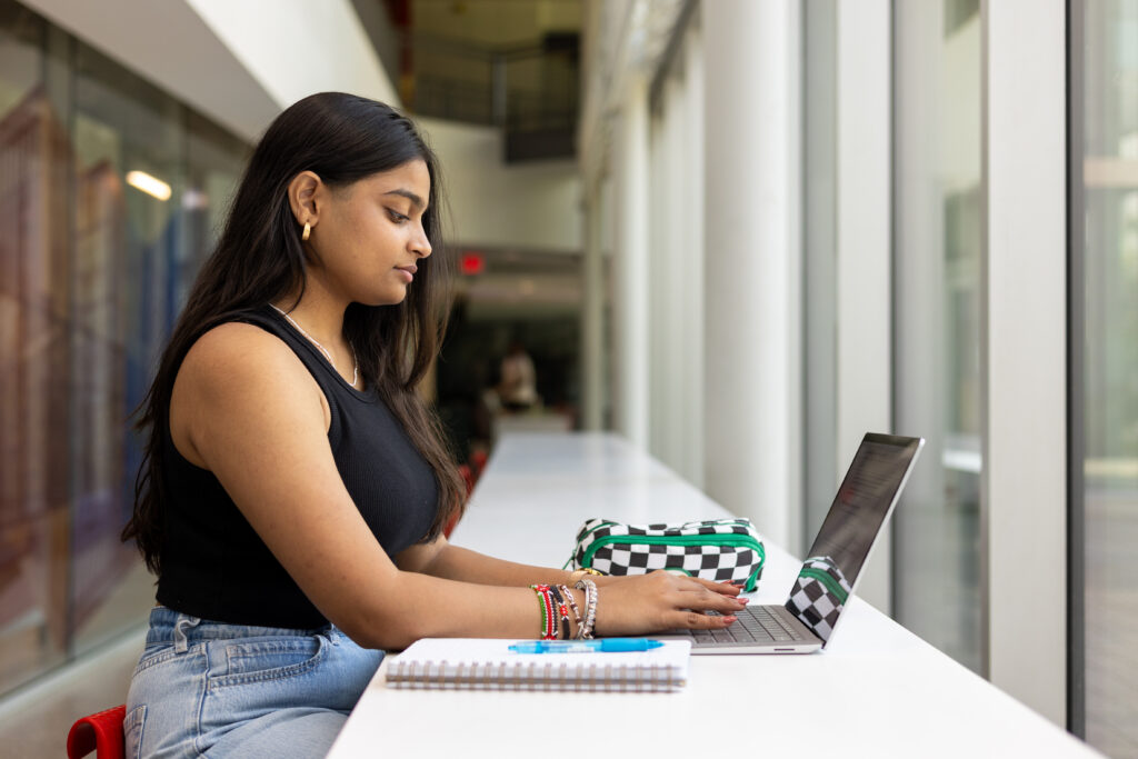 A student writing at their laptop with a notepad and pen beside the laptop. 