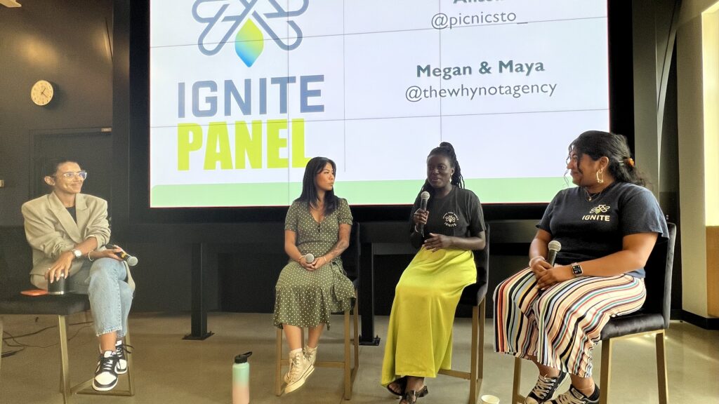 Four women are hosting a panel discussion and sitting on chairs in front of a large screen displaying "IGNITE PANEL.”