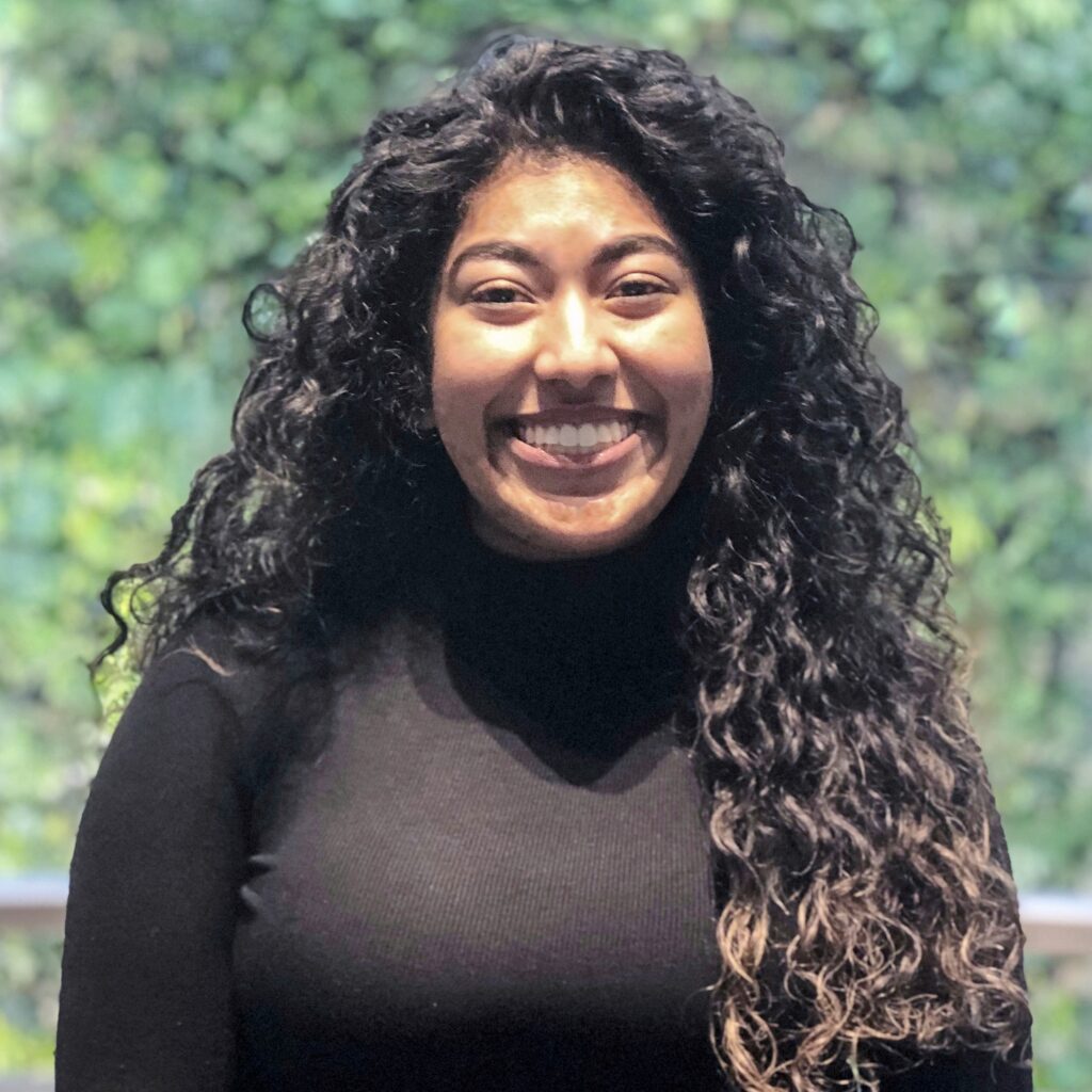 A woman with long, curly dark hair and a black turtleneck is standing in front of a living green wall.