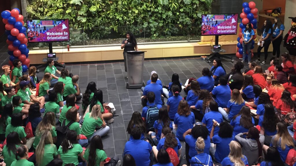 A speaker addresses students wearing colourful shirts at an indoor orientation event. Welcome signs, balloons and a living green wall are visible in the background.