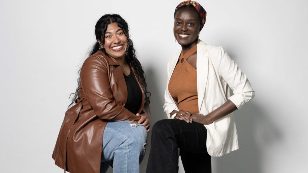 Two women are posing together against a plain background.