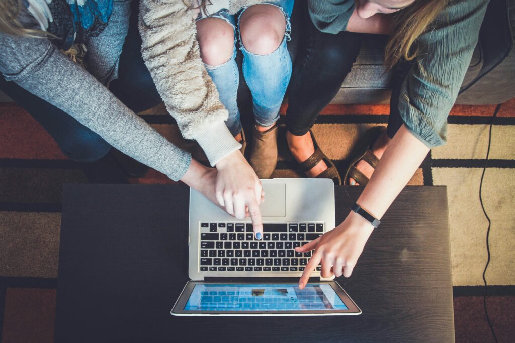 Three people sit around a table, each pointing at a laptop screen in front of them, collaborating on something displayed on the computer.