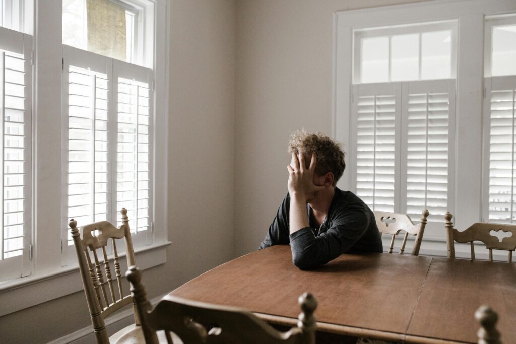 A person ruminating. Sitting at a dinning room table looking out the window with his hand on his face.