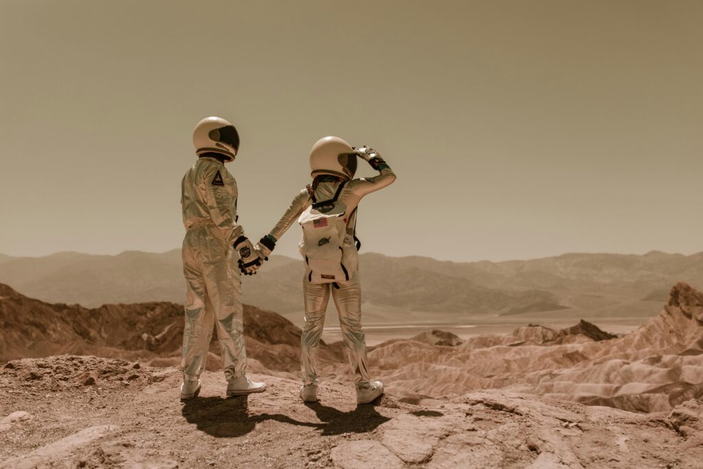 Two people in astronaut gear on a desert plain holding hands.
