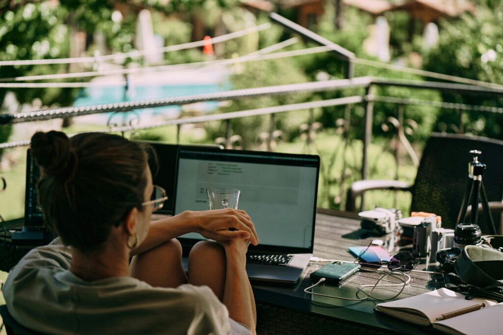 A person sits outdoors at a table with a laptop, smartphone, camera and notebook, surrounded by greenery and blurred background buildings.