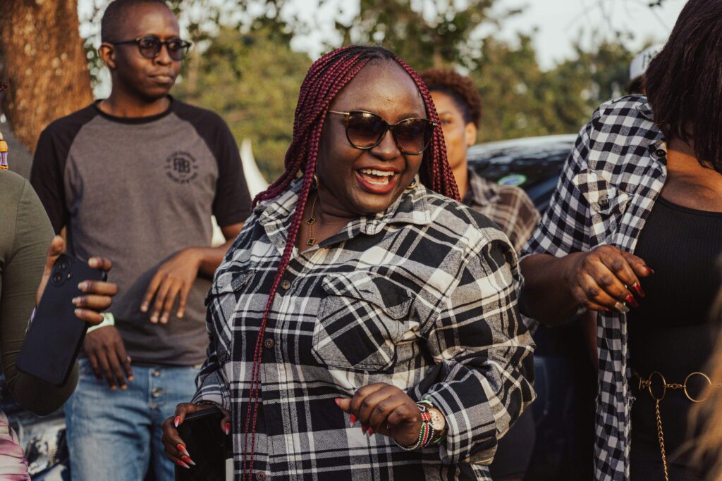 A group of individuals enjoy an outdoor gathering, laughing and dancing. The woman in the centre is wearing sunglasses and a plaid shirt.