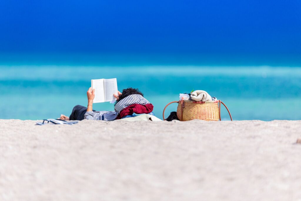 A person is lying on a sandy beach reading a book, with a woven bag and belongings beside them.
