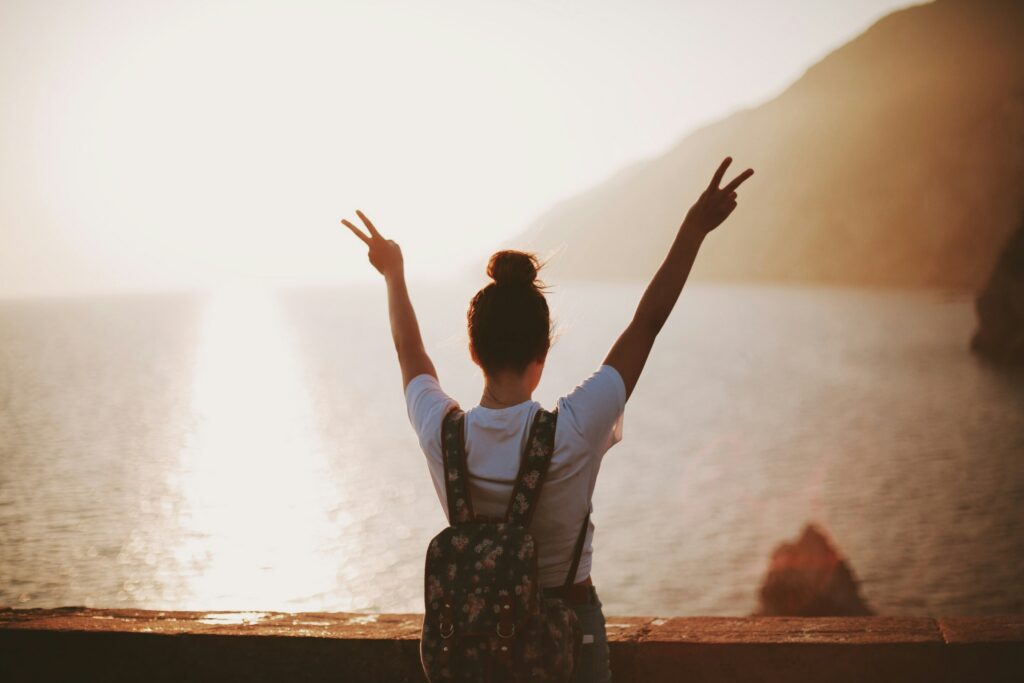 A person with a backpack is sitting on a seaside ledge at sunset, arms raised in a peace sign.